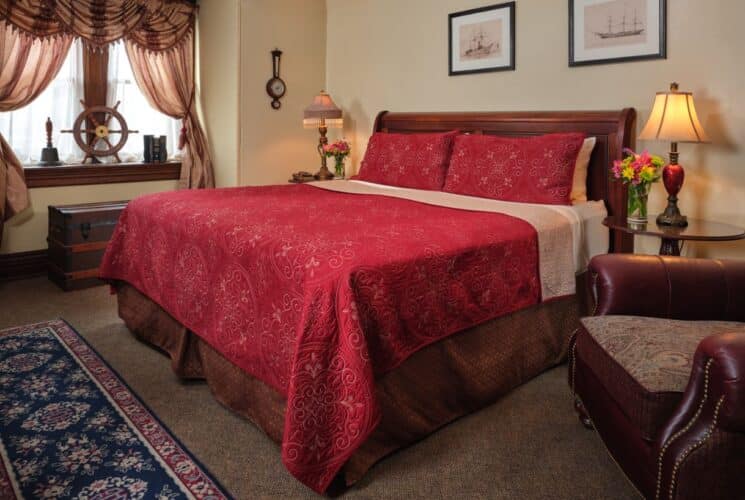 Bedroom with cream walls, dark brown trim, wooden sleigh bed, burgundy bedding, window nook, wood side tables w antique lamps, and and overstuffed chair