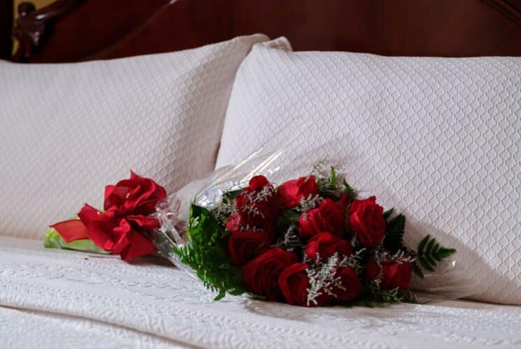 Close up of a bouquet of a dozen red roses laying a a white bed in front of white pillows