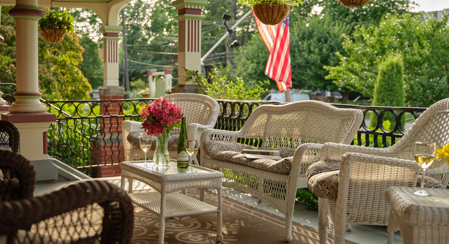 Porch at Spencer House in Erie, Pa. with white and brown wicker furniture, a wicker table with flowers, a bottle of wine glasses on it and an American flag hanging from a porch post in the background