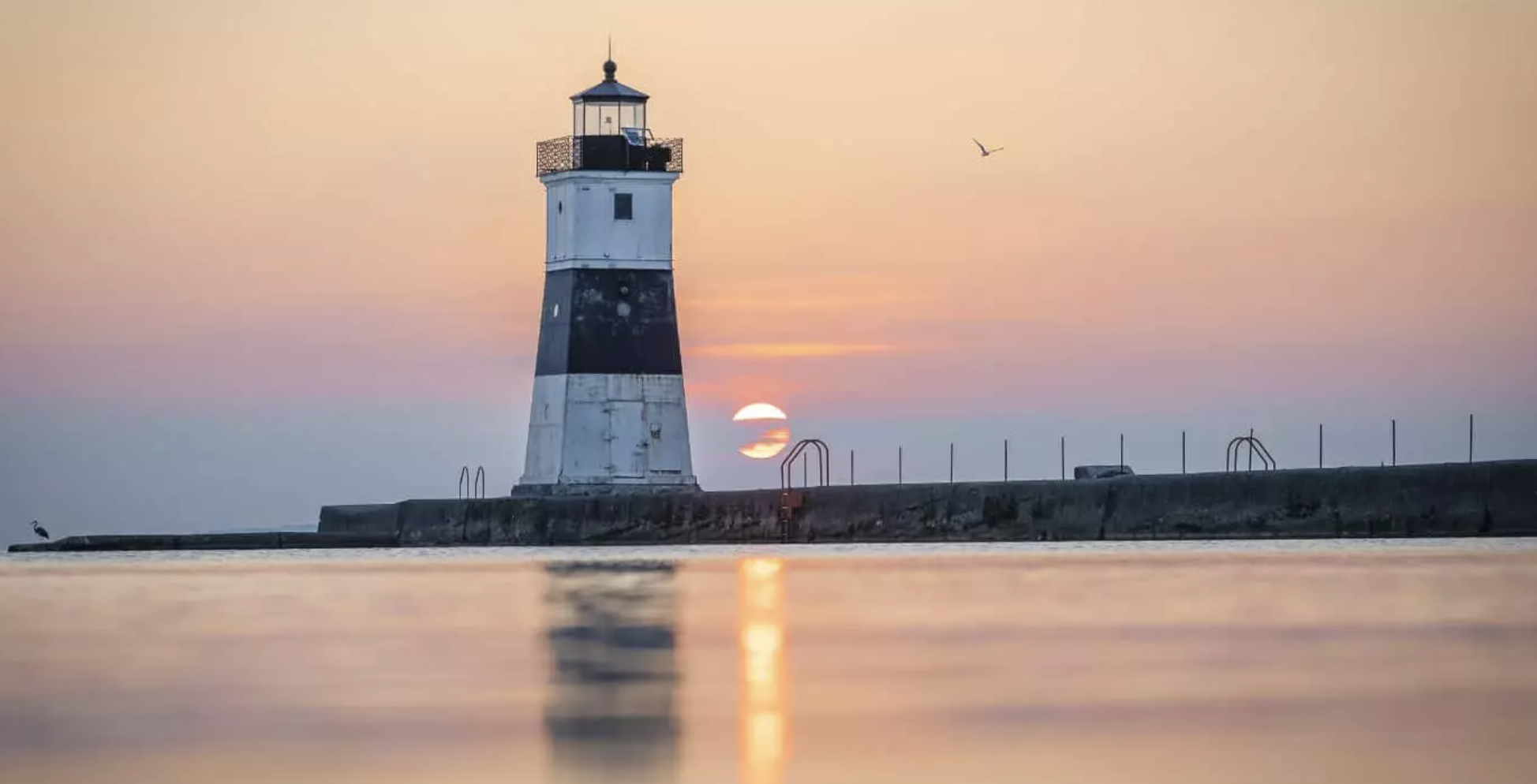 The black and white square North Pier Lighthouse at Presque Isle State Park during a golden sunset, with the sun reflecting on the calm water of Lake Erie.