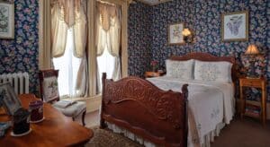 Bedroom with floral wallpaper, tan carpeting, wooden ornate bed, white bedding, wooden nightstands, and large draped windows
