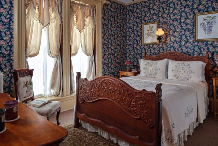 Bedroom with floral wallpaper, tan carpeting, wooden ornate bed, white bedding, wooden nightstands, and large draped windows