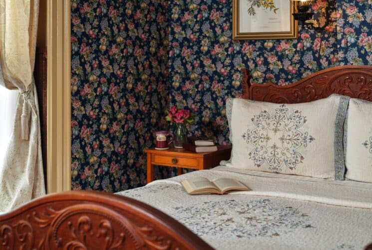Bedroom with floral wallpaper, tan carpeting, wooden ornate bed, white bedding, wooden nightstand, and wall-mounted TV