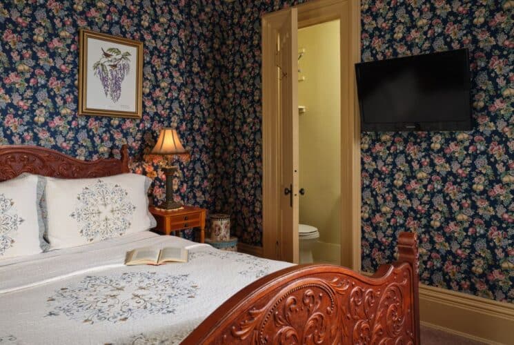 Bedroom with floral wallpaper, tan carpeting, wooden ornate bed, white bedding, wooden nightstands, and wall-mounted TV