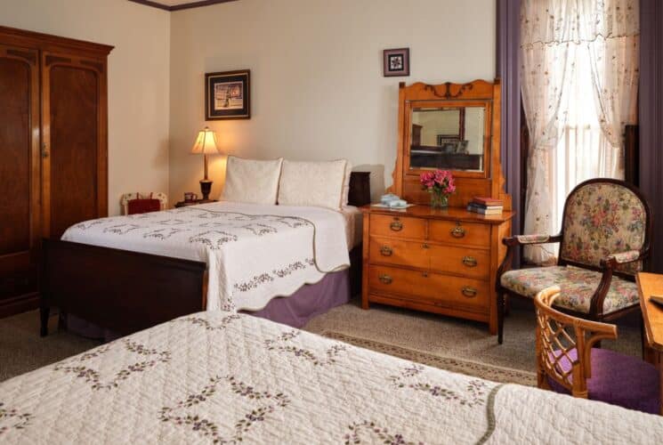 Large bedroom with white walls, two dark wooden beds with multicolored bedding, antique wooden dresser, and wooden armoire