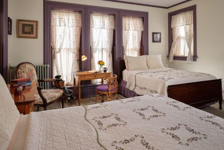 Large bedroom with creme walls, purple trim, carpeting, two dark wooden beds with multicolored bedding, a dresser, a desk and a chair