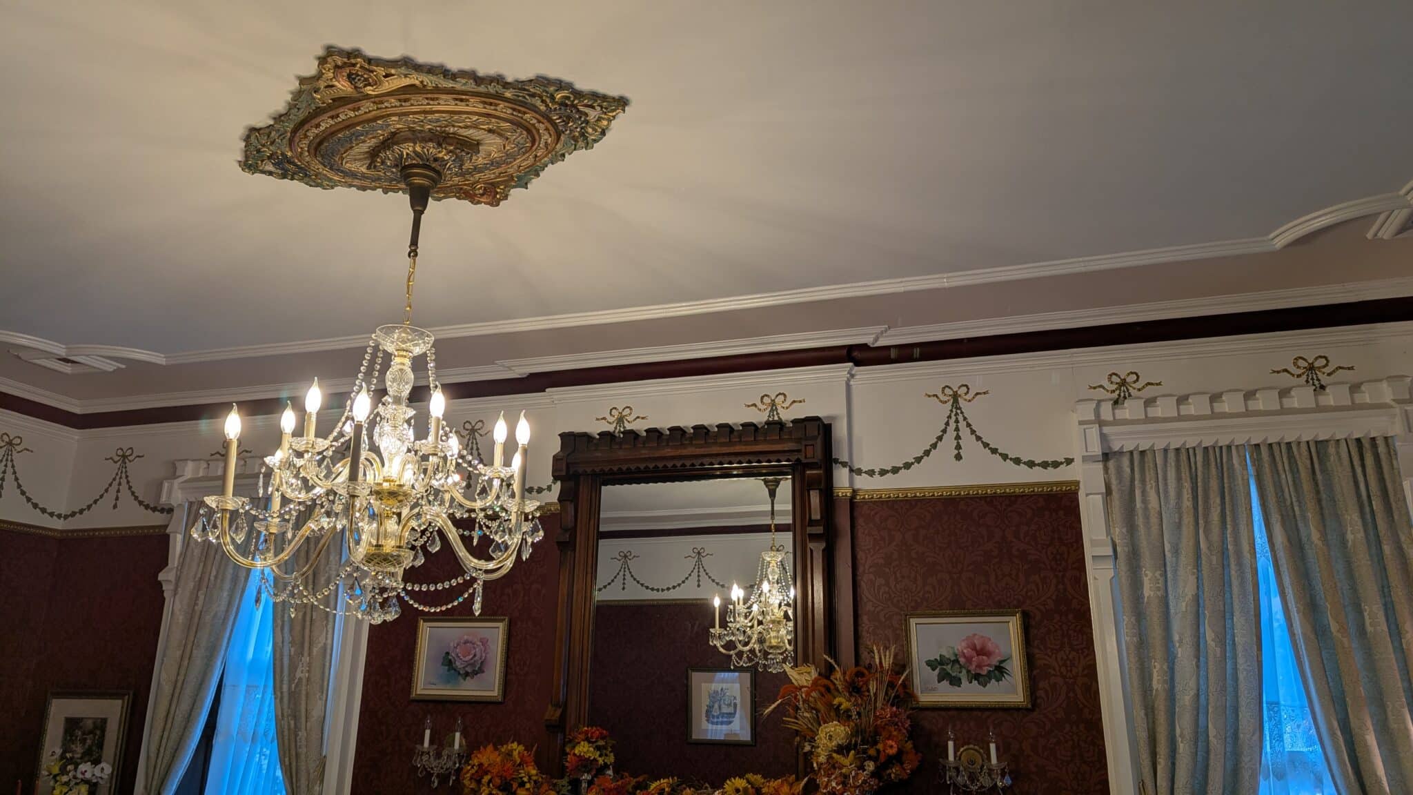 Spencer House parlor fireplace with a tall carved wood mirror frame above the mantel, an ornate plaster ceiling medallion and crystal chandelier, garland and bow plasterwork along the cornice, and a stenciled damask-pattern paint finish on the burgundy walls.