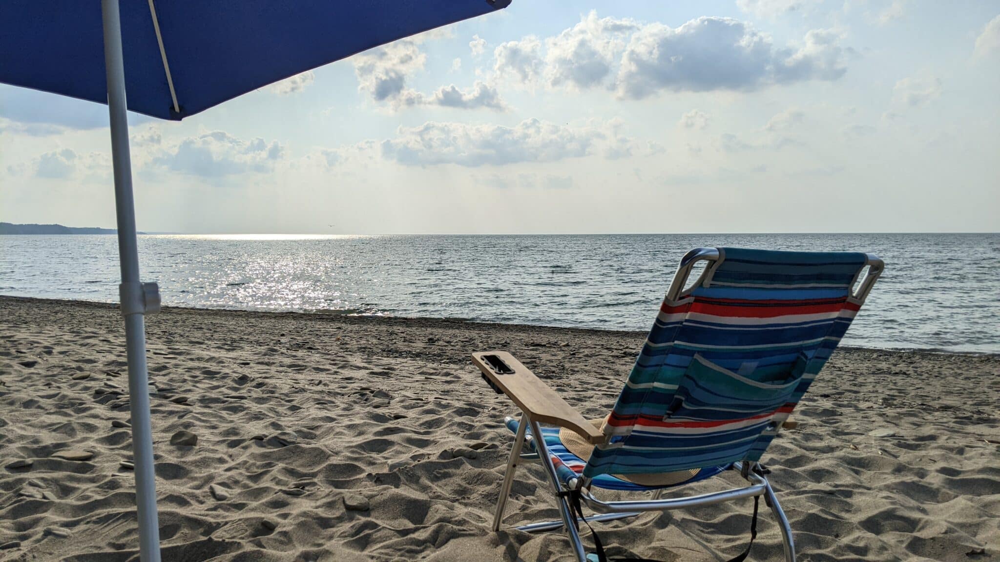Photo of a Presque Isle State Park beach looking out onto Lake Erie with a blue beach umbrella, a beach chair with multi color stripes and a beach hat on the chair.
