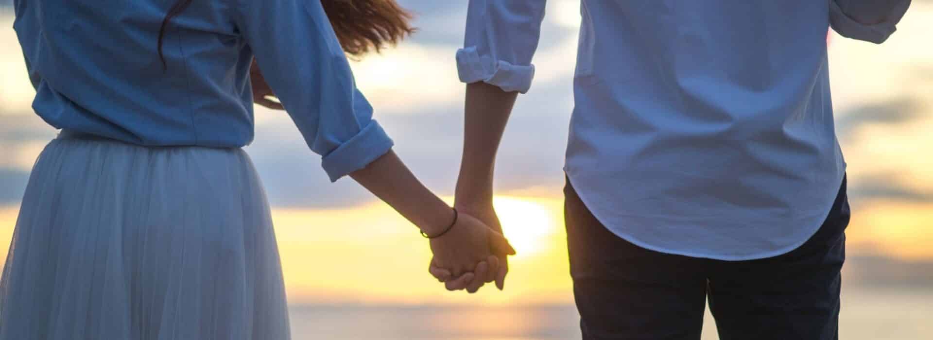 Photo form behind of two people holding hands in front of a sunset