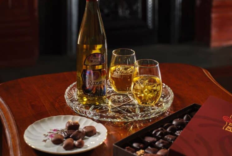 Close up view of a box of chocolates, a small plate with chocolates, partially filled wine glasses, and a bottle of white wine on a wooden coffee table