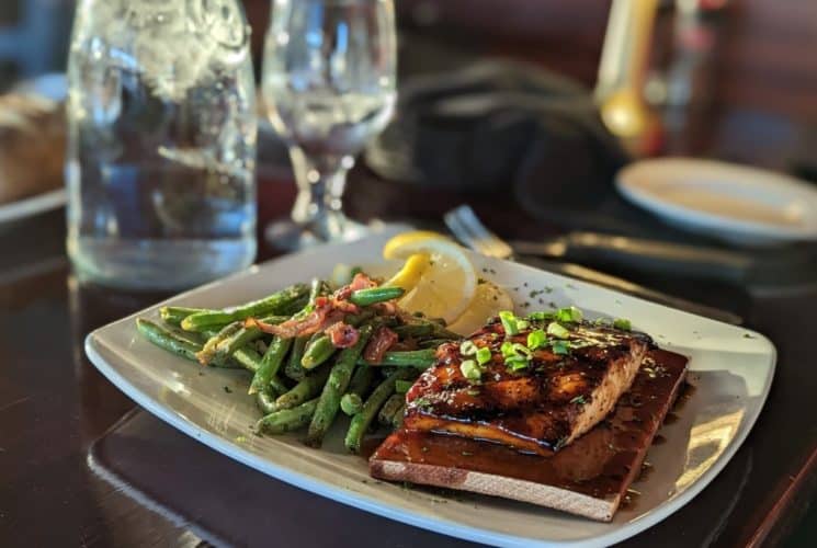 White plate with salmon on cedar blank, bacon-topped green beans, and slices of lemon on a wooden table