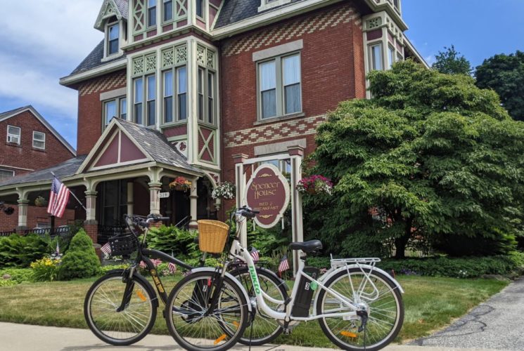 Red brick house with white trim, front porch, large Spencer House Bed & Breakfast sign, surrounded by lush green trees, shrubs, plants, and grass, and two electric bikes on the sidewalk