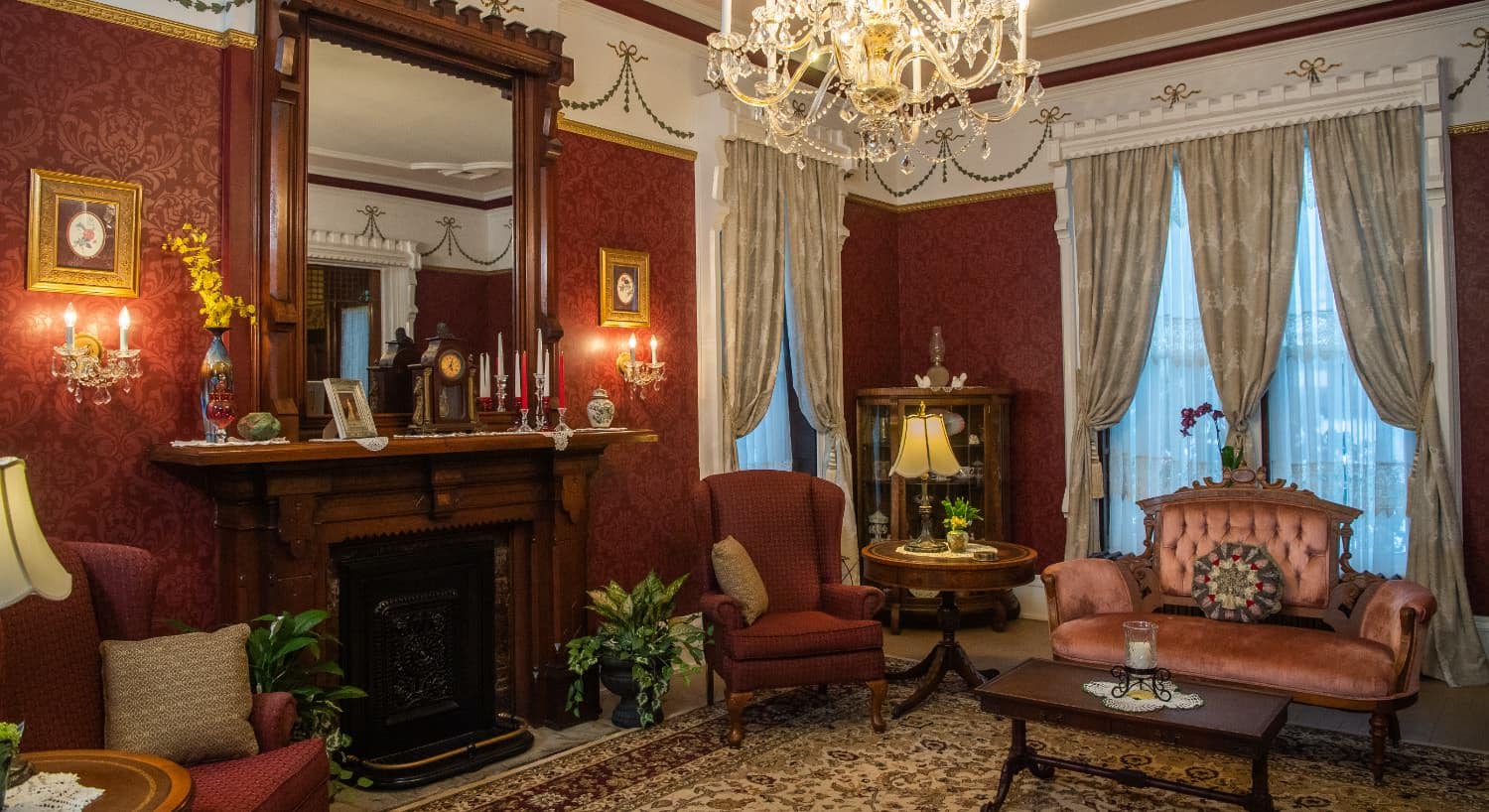 Large room with ornate burgundy wall paper, tall ceilings, large windows with drapery, red upholstered armchairs, and antique fireplace with wooden mantel and large mirror