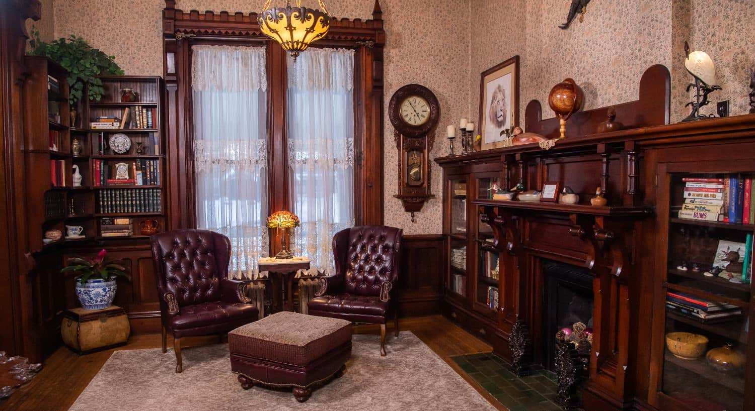 Study with dark wooden cabinets, wooden fireplace, two leather armchairs and ottoman, and large windows