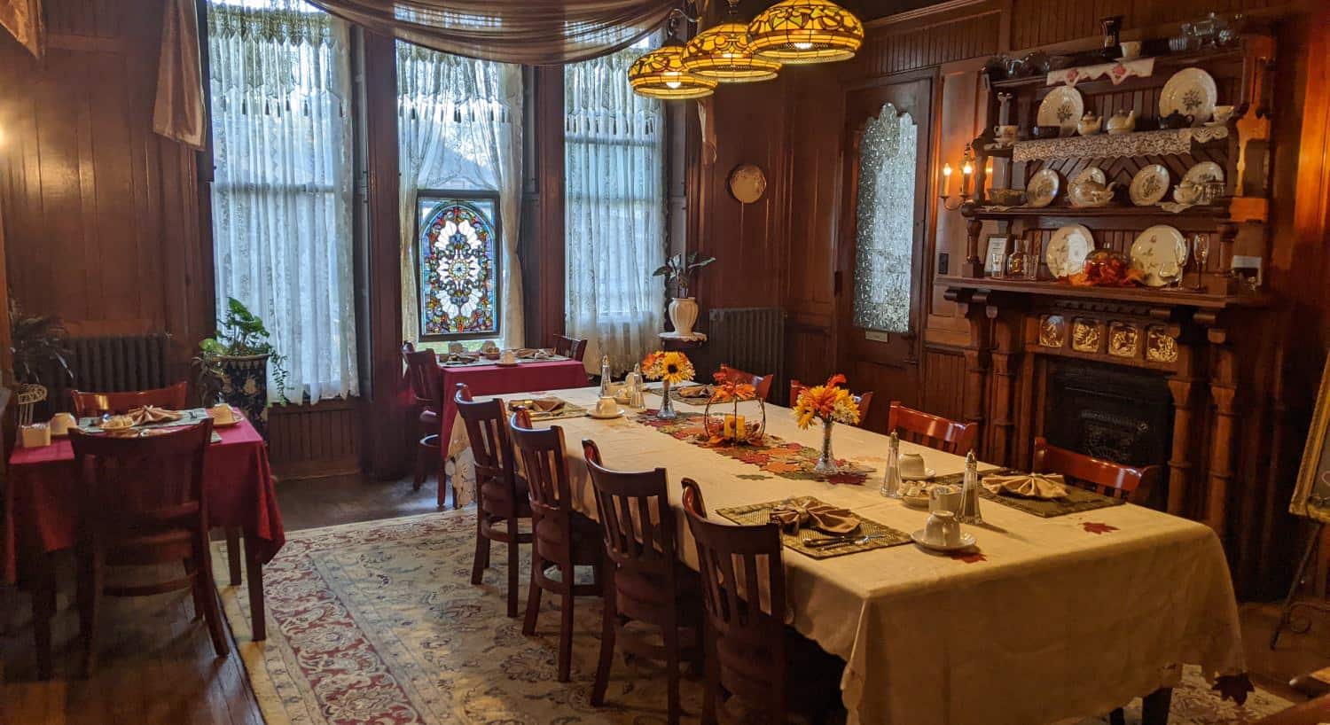 Large dining room with wooden paneling and flooring, fireplace with wood mantel, two small dining tables, and one large dining table with multiple placesettings