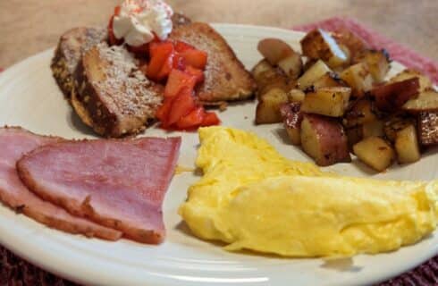 white plate with scrambled eggs, ham, french toast and seasoned potatoes