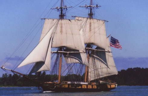 Large schooner ship with white sails and a United States Flag on the water with trees in the background