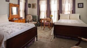 Large bedroom with white walls, gray trim, two dark wooden beds with multicolored bedding, antique wooden dresser, chair and small desk