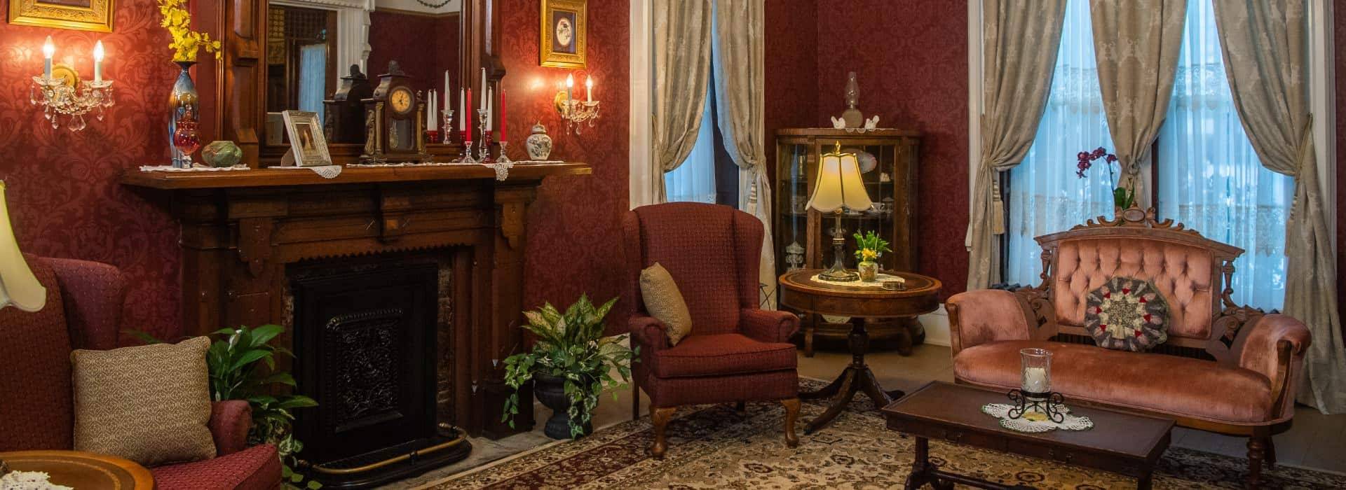 Large gathering room with burgundy wallpaper, vintage wooden fireplace, China cabinet, red upholstered arm chairs, and pink upholstered antique love seat